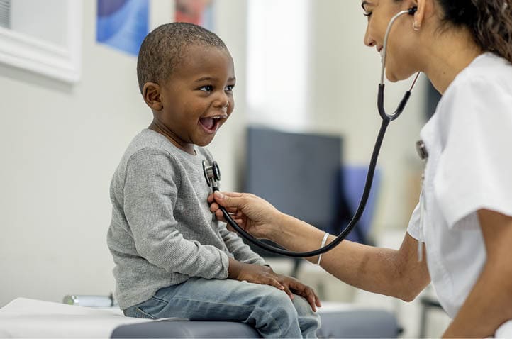 A young boy of African decent, sits up on an exam table as a female Paediatrician preforms a check-up on him. The boy is dressed casually and smiling as the doctor listens to his heart.
